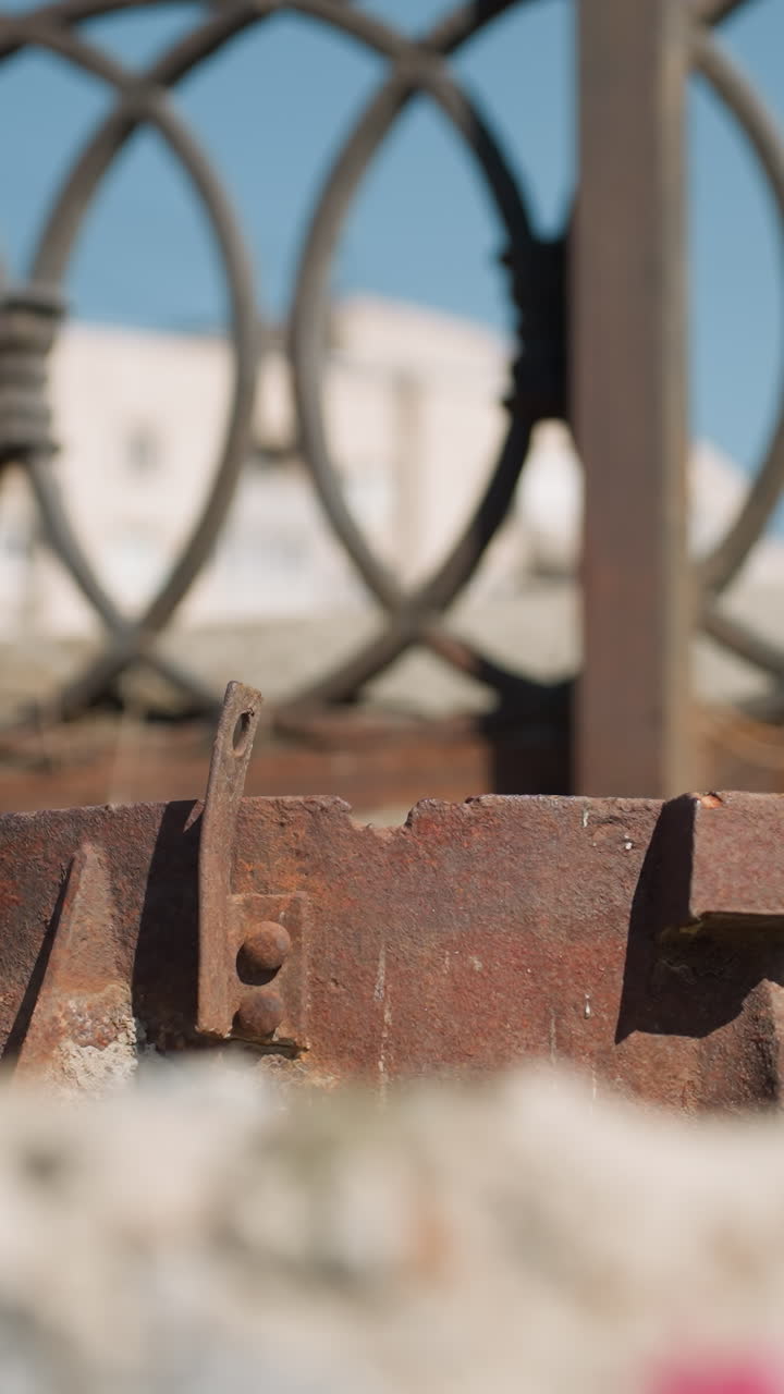vista de cerca de un objeto metálico oxidado en el suelo con una valla ornamentada y edificios urbanos en el fondo, con un paso por