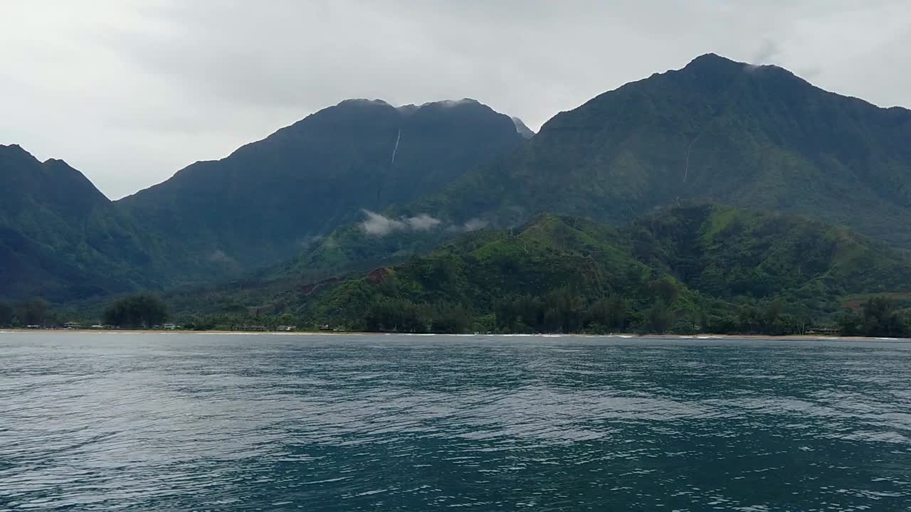 hd 120fps 하와이 카우아이는 전경에 보트 스프레이가 있는 흐린 거리에 폭포가 있는 녹색 언덕과 산이 있는 오른쪽에서 왼쪽으로 떠 있는 바다에서 보트 타기