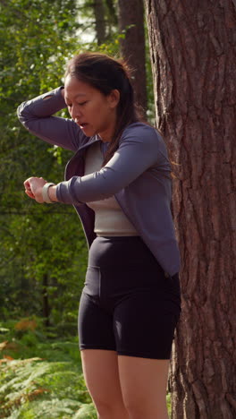 Video-Vertical-De-Una-Mujer-Haciendo-Ejercicio-Al-Aire-Libre-Corriendo-Por-Una-Pista-En-El-Bosque,-Deteniéndose-Para-Tomar-Aire-Y-Verificando-La-Aplicación-De-Actividad-En-Un-Reloj-Inteligente,-Filmado-En-Tiempo-Real-1