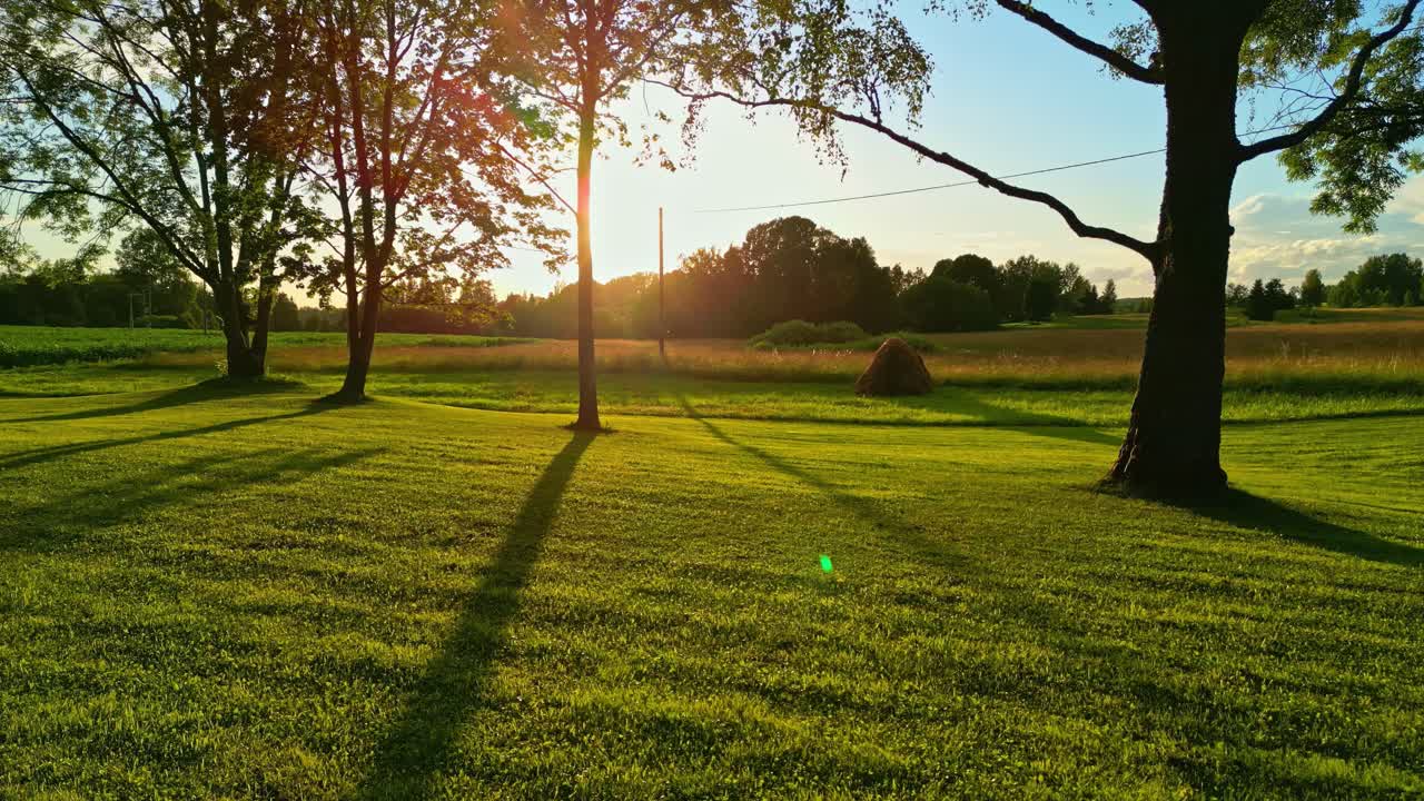 Golden sunset casting long tree shadows across green meadow with haystack and rural fields
