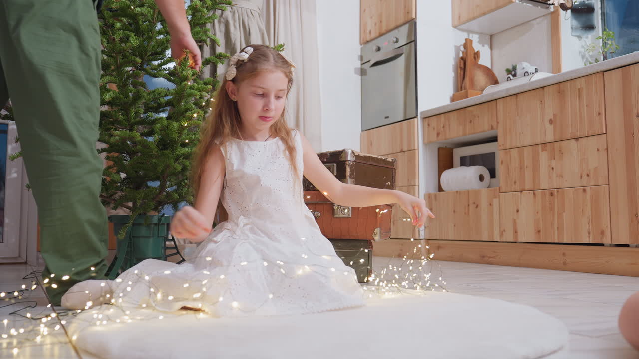 Little girl in white gown sitting on white rug stretching out Christmas light in preparation for decorating, smiling in festive spirit, helping with holiday tree decorating at home