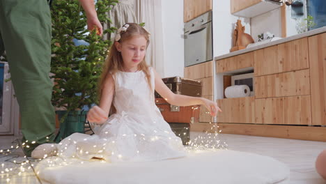 little girl in white gown sitting on white rug stretching out christmas light in preparation for decorating, smiling in festive spirit, helping with holiday tree decorating at home