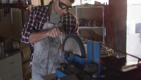 caucasian male knife maker in workshop wearing glasses and using sander