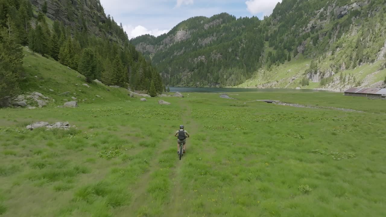 toma de seguimiento de drones de un ciclista masculino montando una bicicleta de montaña en un camino de hierba entre montañas durante el verano