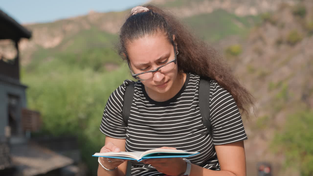 Studente concentrato che legge un quaderno all'aperto vicino a una collina, intensa concentrazione con zaino e occhiali, sfoglia le pagine mentre studia vicino a edifici di un villaggio rustico, luce solare calda e ritratto spontaneo