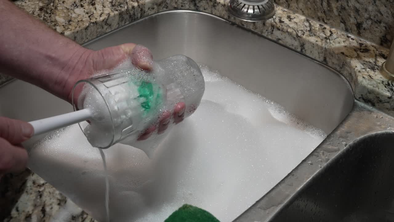 A man scrubs a cup in a foam bubble-filled stainless steel sink
