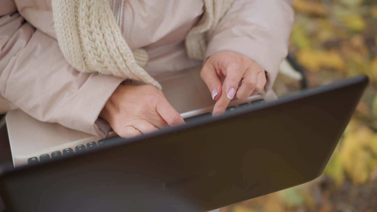 top down close up of woman typing on laptop while seated outdoors on park bench in autumn, hands with painted nails moving across keys against backdrop of colorful leaves