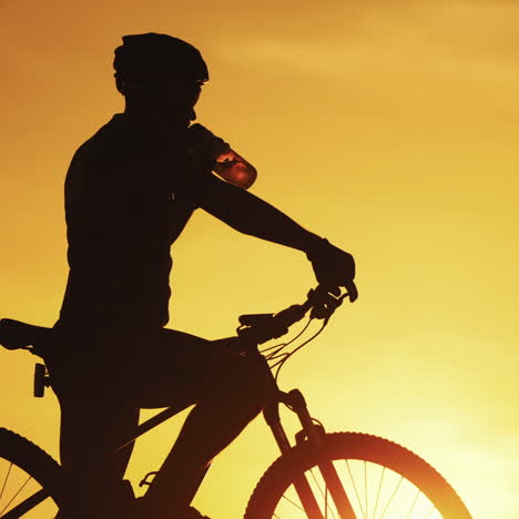 silhouette of professional bicyclist taking a bottle and drinking water at golden sunset. male cyclist in helmet drinks water on the natural background of setting the sun in the evening.