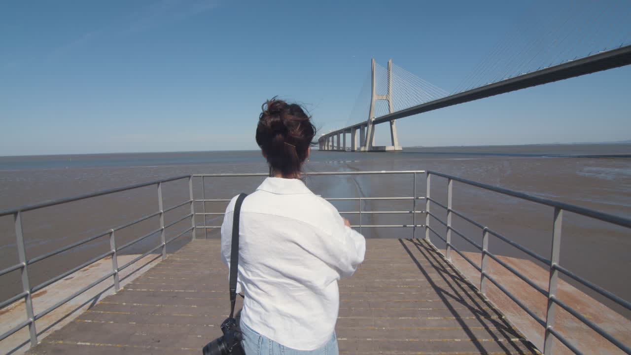 Follow shot of a young woman walking down the pier and enjoying the amazing view of the river with a bridge. Low tide.Tagus river. Contemplation in solitude. Architectural wonder