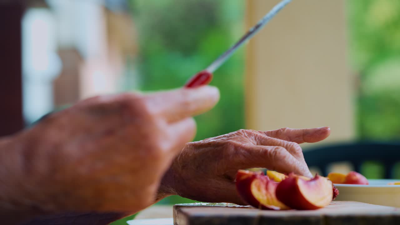 Senior woman slices a fresh peach with a knife on a rustic table outdoors surrounded by greenery