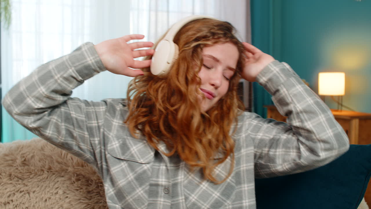 Woman sitting on couch wearing headphones dancing with closed eyes enjoying favorite relaxing music