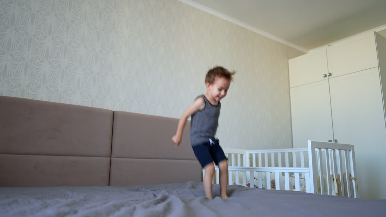 Brunet Caucasian toddler boy lies on the double bed. Kid stands up and starts to jump happily. Low angle view.