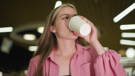 lady in pink dress seated in a restaurant holds her coffee cup and takes a sip with a satisfied expression, enjoying the taste, the background features a bokeh light effect and blurred view of people