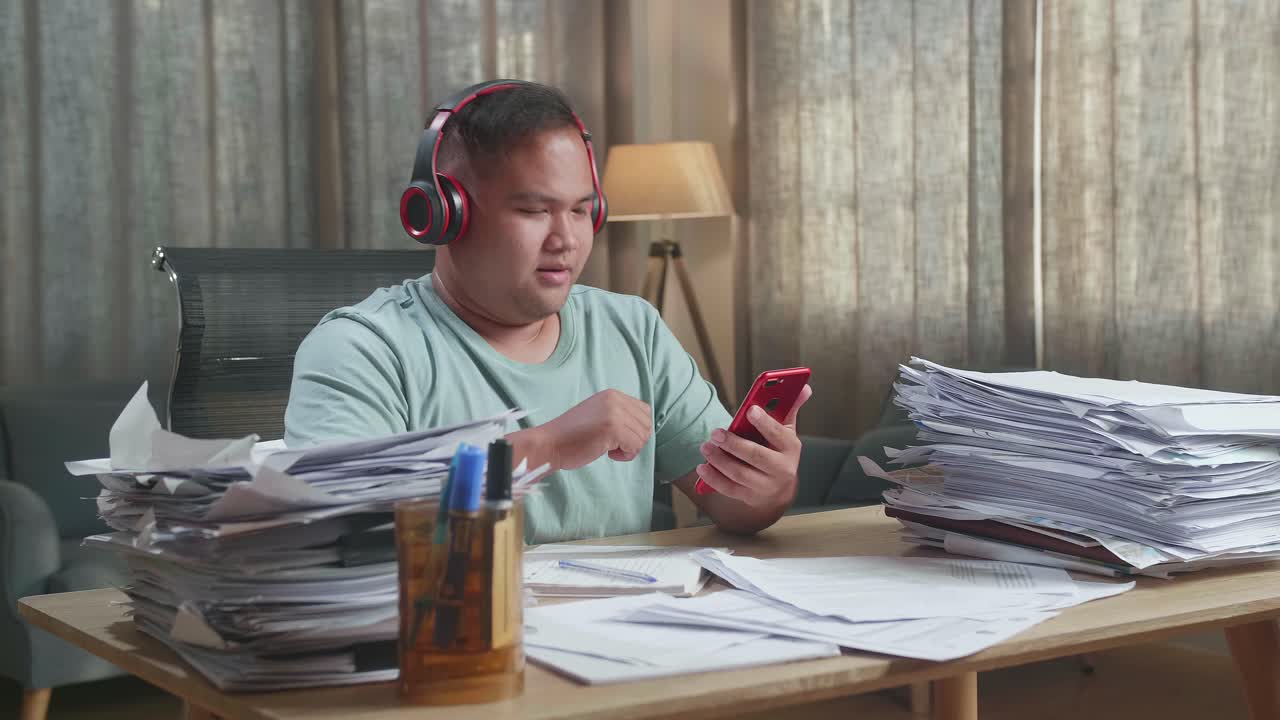 Fat Asian Man Wearing Headphones And Listening To Music On Smartphone After Working With Documents At The Office