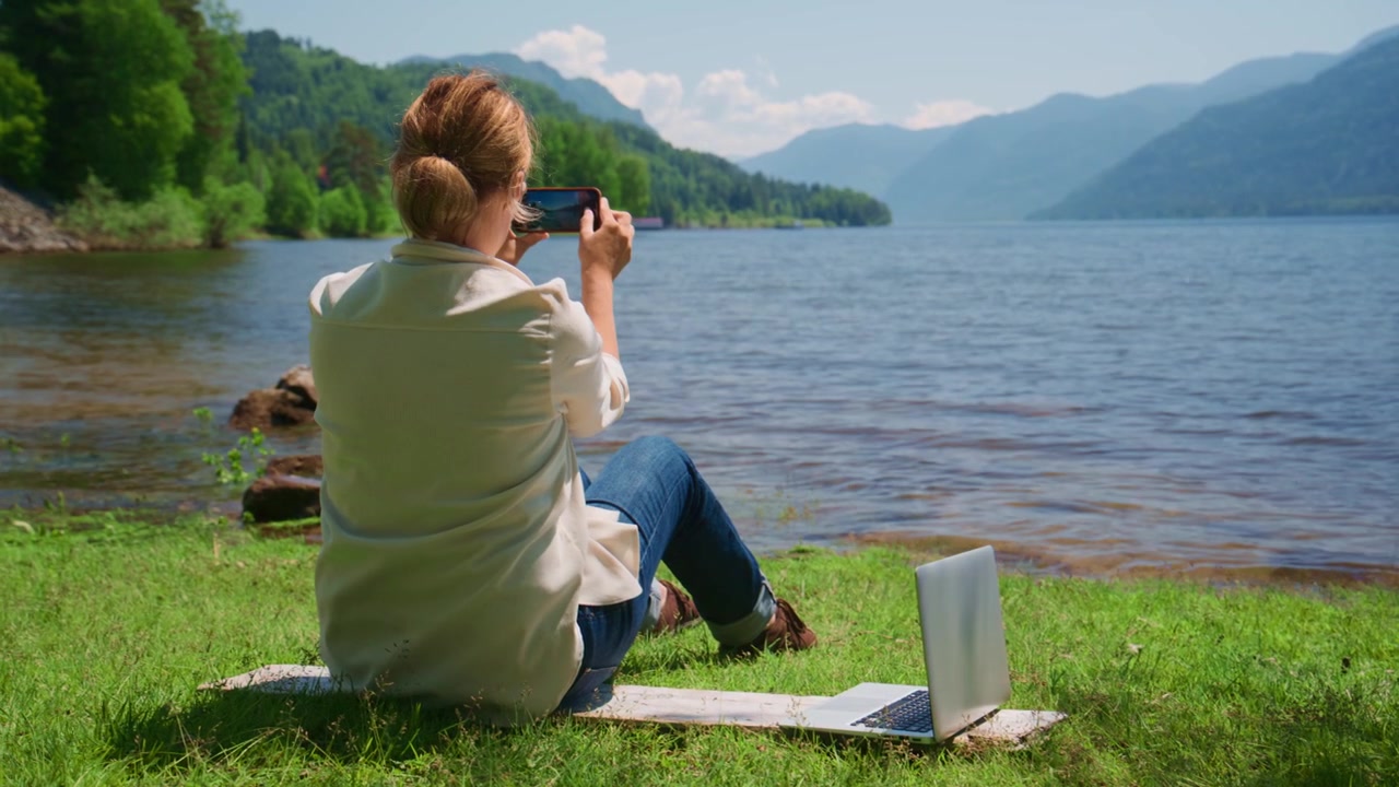Woman sits by a serene lake, capturing its beauty with her phone. She relaxes with a laptop open beside her, enjoying the view. Mountains rise behind the calm water, lush with green trees