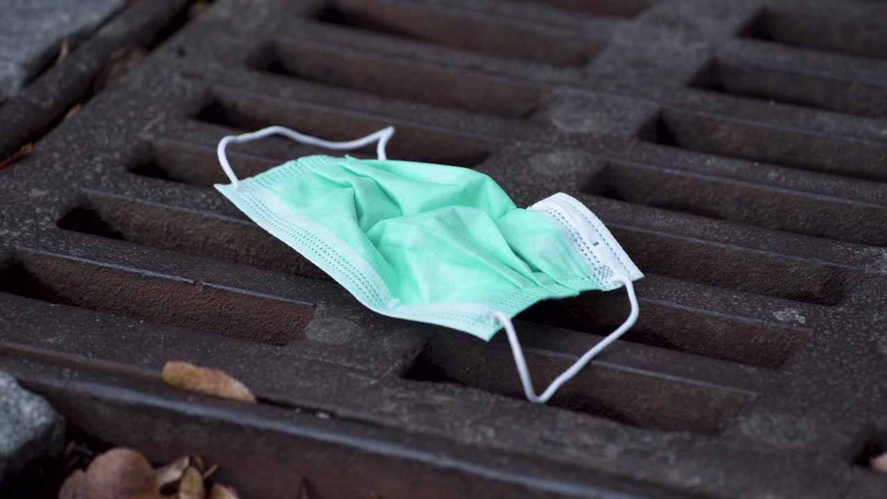 단순한 녹색 보호용 covid-19 안면 마스크가 마른 나뭇잎 사이에 있는 직사각형의 녹슨 하수도 해치에 버려져 있고, 지나가는 사람들, 거의 밟을 뻔한 것, 정적인 클로즈업 4k 샷