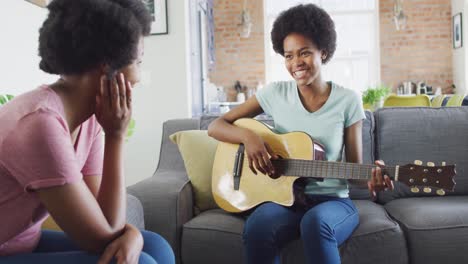 Happy-african-american-mother-and-daughter-sitting-on-sofa-playing-guitar