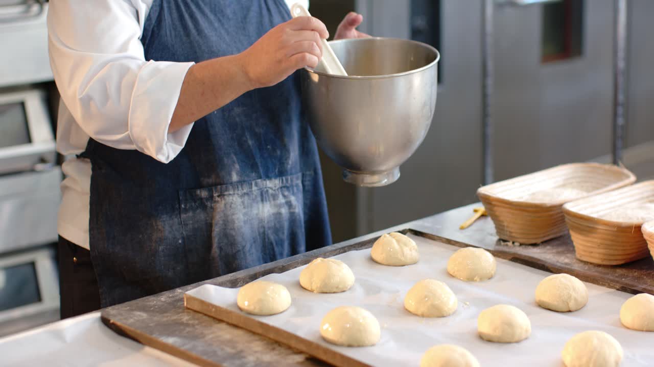 African American woman baker glazing rolls with red-brush at bench dipping into bowl to refill
