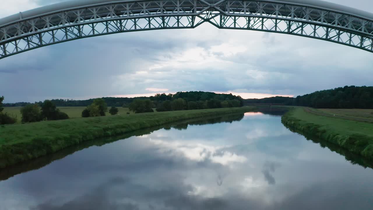 Flying backwards under two bridges for gas pipes running across the calm river Moravia on the border of Slovakia and Czechia. Aerial sunset footage of symmetrical gas pipe bridges reflecting in water