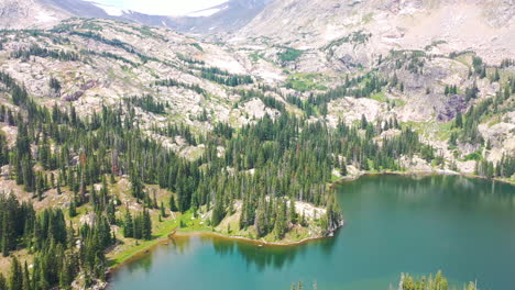 aerial drone tilt up to reveal tall mountain peak next to clear blue water lake surrounded by thick pine tree forest in nederland colorado rocky mountains