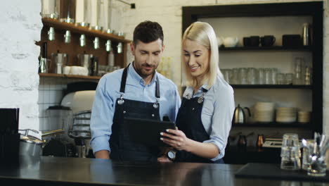 Cheerful-Good-Looking-Waiter-And-Waitress-Standing-At-The-Bar,-Talking-And-Watching-Some-Video-On-The-Tablet-Computer