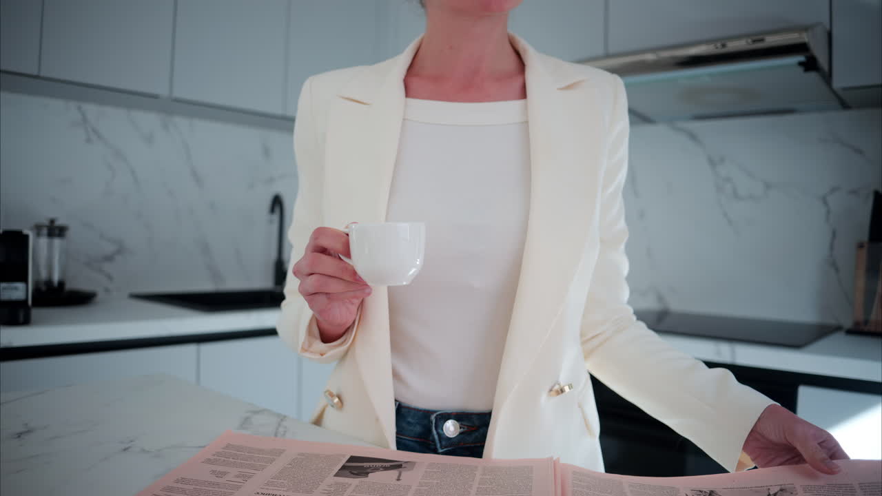 Woman in a white blazer sipping coffee while reading the newspaper in the kitchen