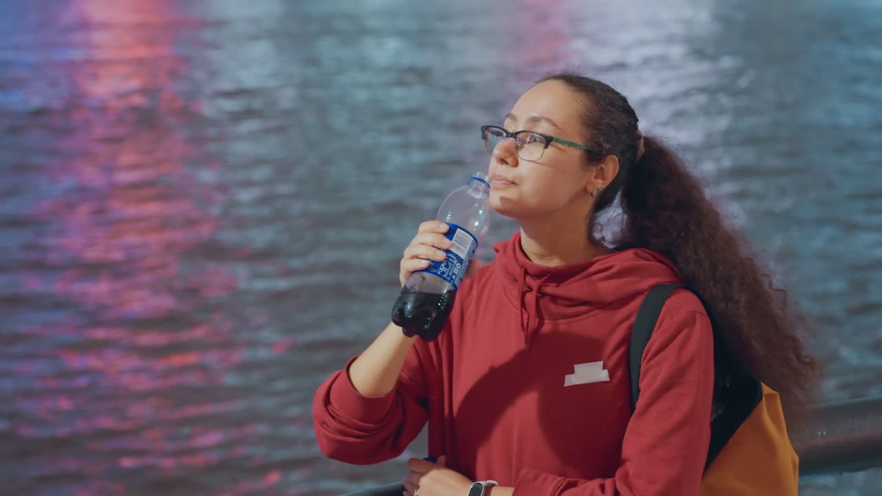 Giovane studentessa che beve acqua sul lungomare, tenui riflessi al neon sul fiume, felpa e zaino, occhiali e coda di cavallo, sguardo contemplativo, calma atmosfera notturna della città, ritratto spontaneo con sottile