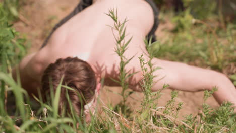 top view of muscular warrior crawling shirtless on dirt hill with arms outstretched, showing center of hair and intense effort, surrounded by dense green vegetation, dry soil, and warm sunshine