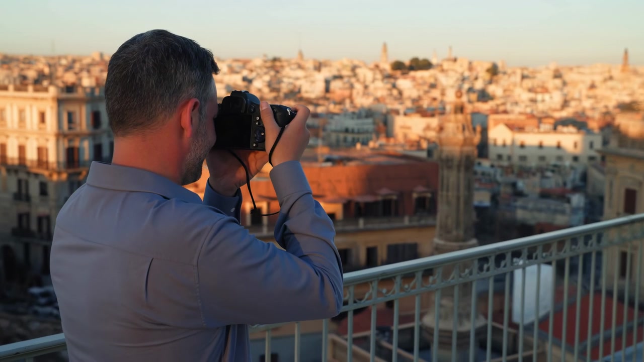 Visto da dietro, un uomo solleva una fotocamera a mano dal livello del petto all'altezza degli occhi mentre si affaccia su un vasto skyline urbano, con la calda luce solare radente che illumina edifici e tetti a strati