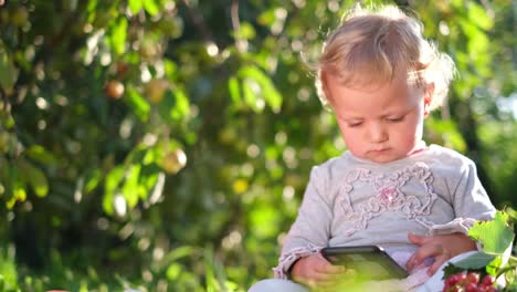 cute little baby holding a smartphone and looking at the screen in nature little girl looks at her phone she is sitting on a blanket in the garden