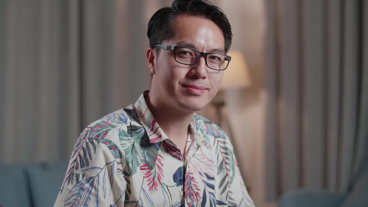 primer plano de un hombre asiático en silla de ruedas sonriendo y cruzando los brazos hacia la cámara mientras edita el video por un escritorio al lado de la cámara en casa