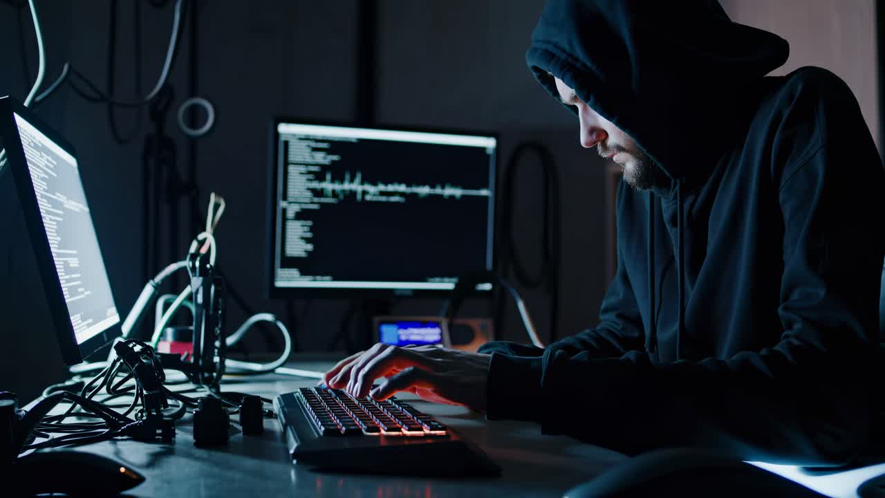 A hooded figure types at a computer in a dimly lit room, captured from a side angle