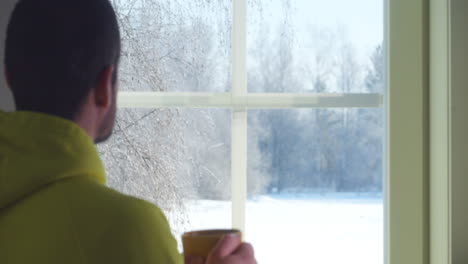 man in hoodie sips coffee, looks outside at winter scenery