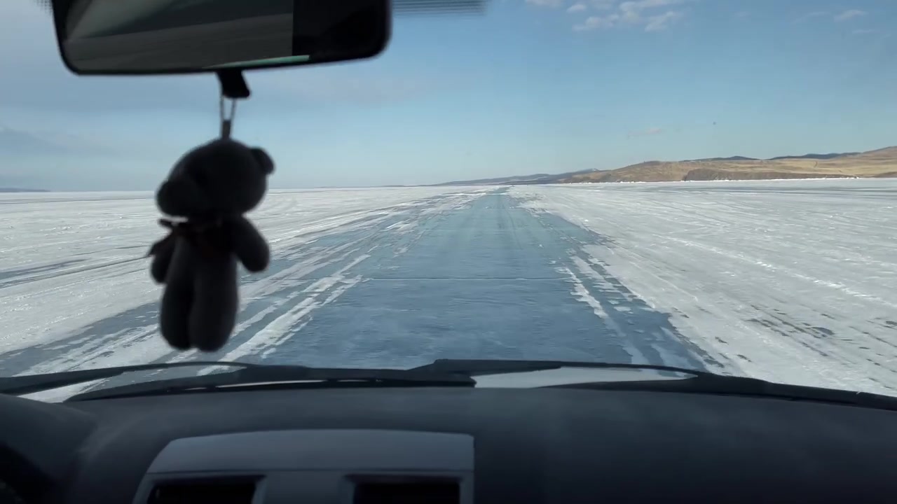 POV dall'interno di un'auto che percorre una strada di ghiaccio attraverso il lago Baikal ghiacciato in inverno, con cielo azzurro limpido e colline distanti, Siberia, Russia.