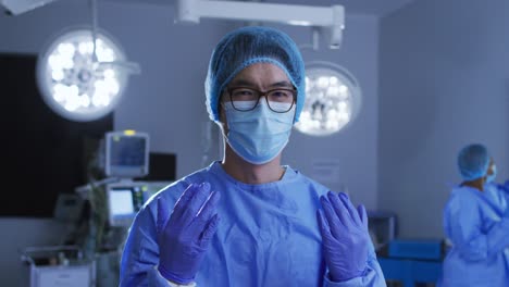 portrait of asian male surgeon wearing face mask, gloves, cap and scrubs in operating theatre