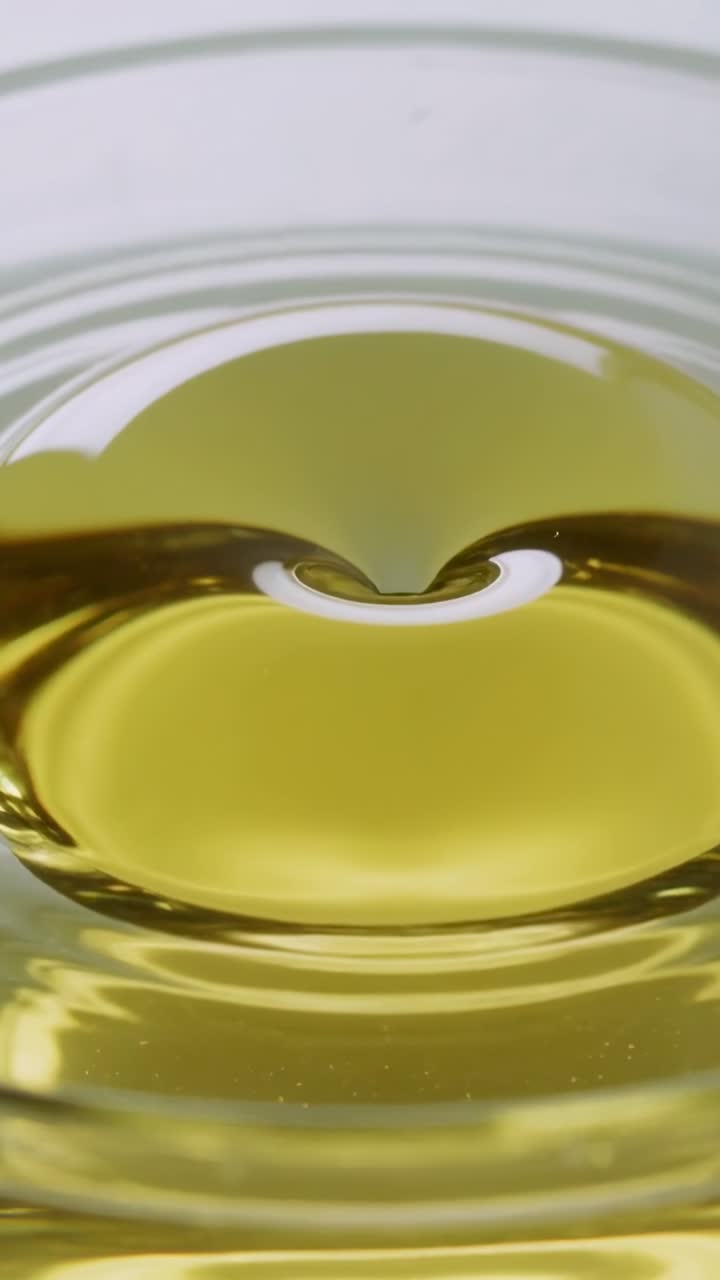 Video verticale: Gocciolina che impatta creando una cavità e increspature in olio giallo all'interno di una ciotola di vetro.