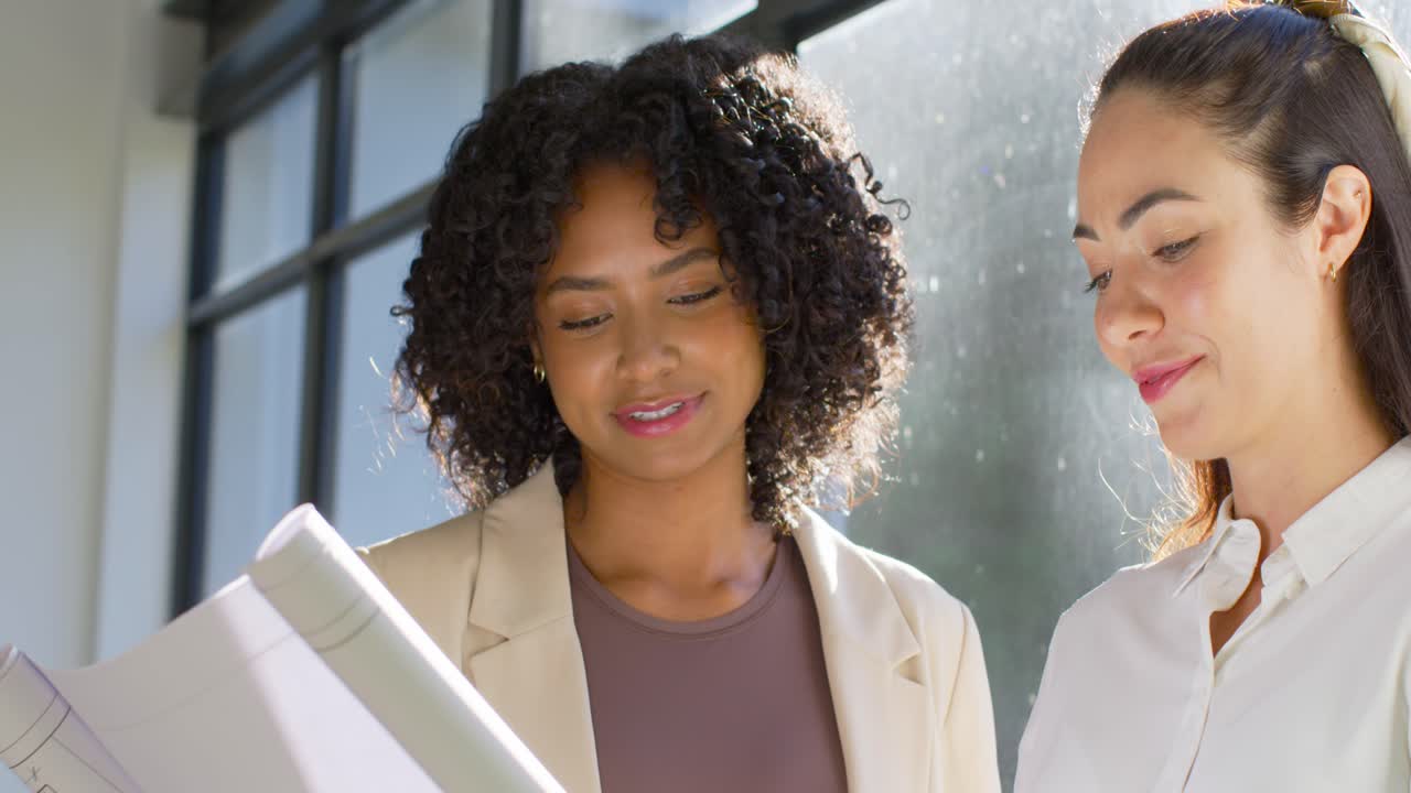 Colegas de trabalho femininas diversas vestindo trajes de negócios em uma janela com grade, inclinando uma pasta e revisando páginas.