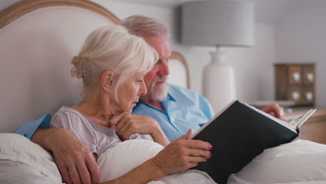 Retired-Senior-Couple-In-Bed-At-Home-Looking-At-Photo-Album-Together