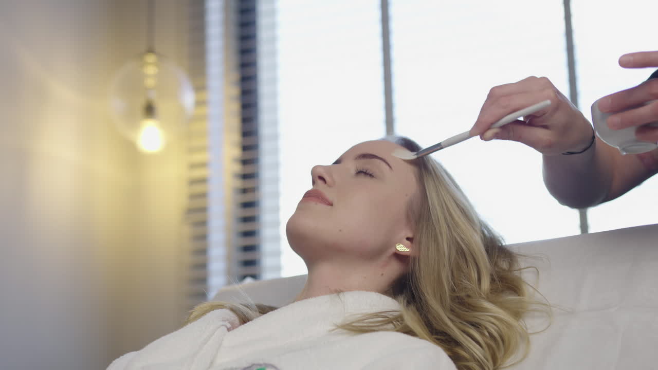 Eine professionelle Kosmetikerin trägt mit einem Pinsel eine kosmetische Creme aus natürlichen Kräuterölen auf das Gesicht einer blonden Patientin in einem modernen Luxus-Spa-Salon auf – eine Hautpflegebehandlung.