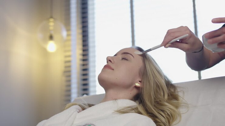 Eine professionelle Kosmetikerin trägt mit einem Pinsel eine kosmetische Creme aus natürlichen Kräuterölen auf das Gesicht einer blonden Patientin in einem modernen Luxus-Spa-Salon auf – eine Hautpflegebehandlung.