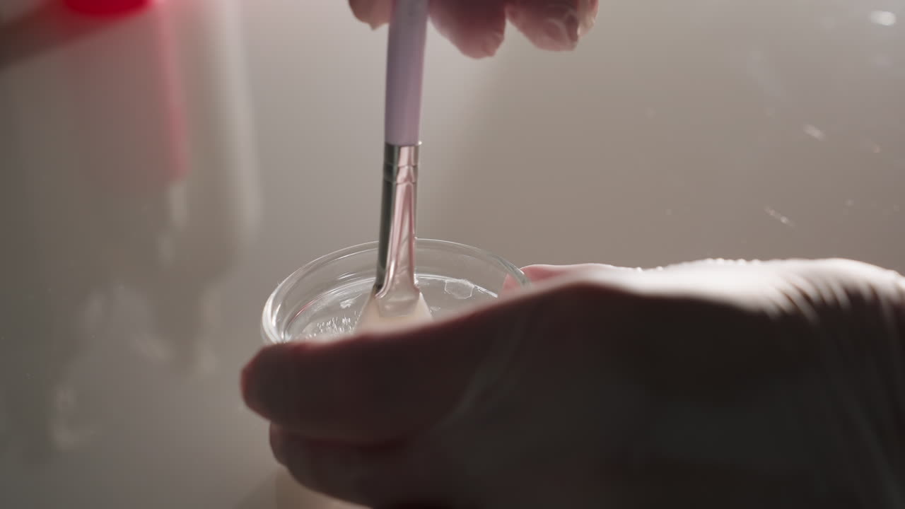 Hands in gloves mix creamy facial mask in small glass bowl with silicone brush on reflective spa trolley under soft light, conveying professional skincare ritual and hygienic precision care