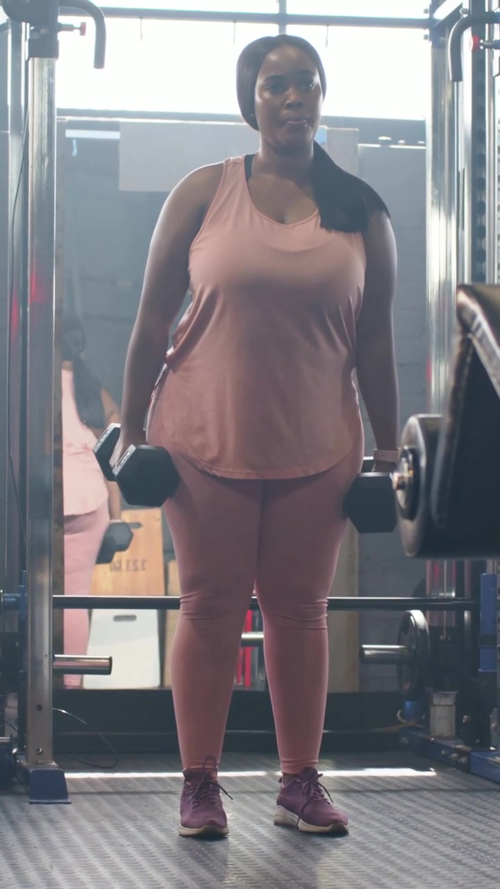 Video verticale: Donna in rosa che inizia una serie sollevando un manubrio esagonale destro vicino allo specchio della palestra per la forza.