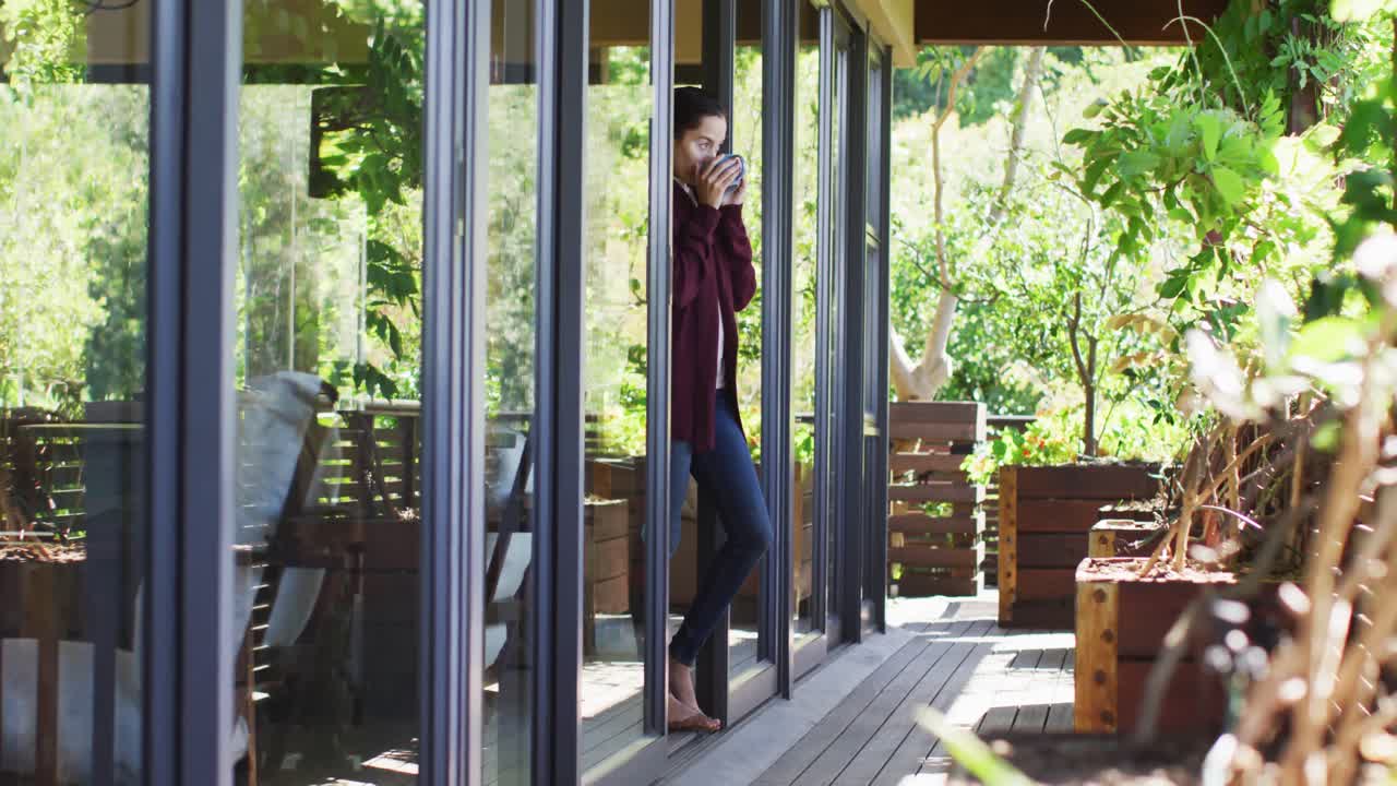 mujer biracial relajada bebiendo café, de pie en el patio, mirando a la distancia