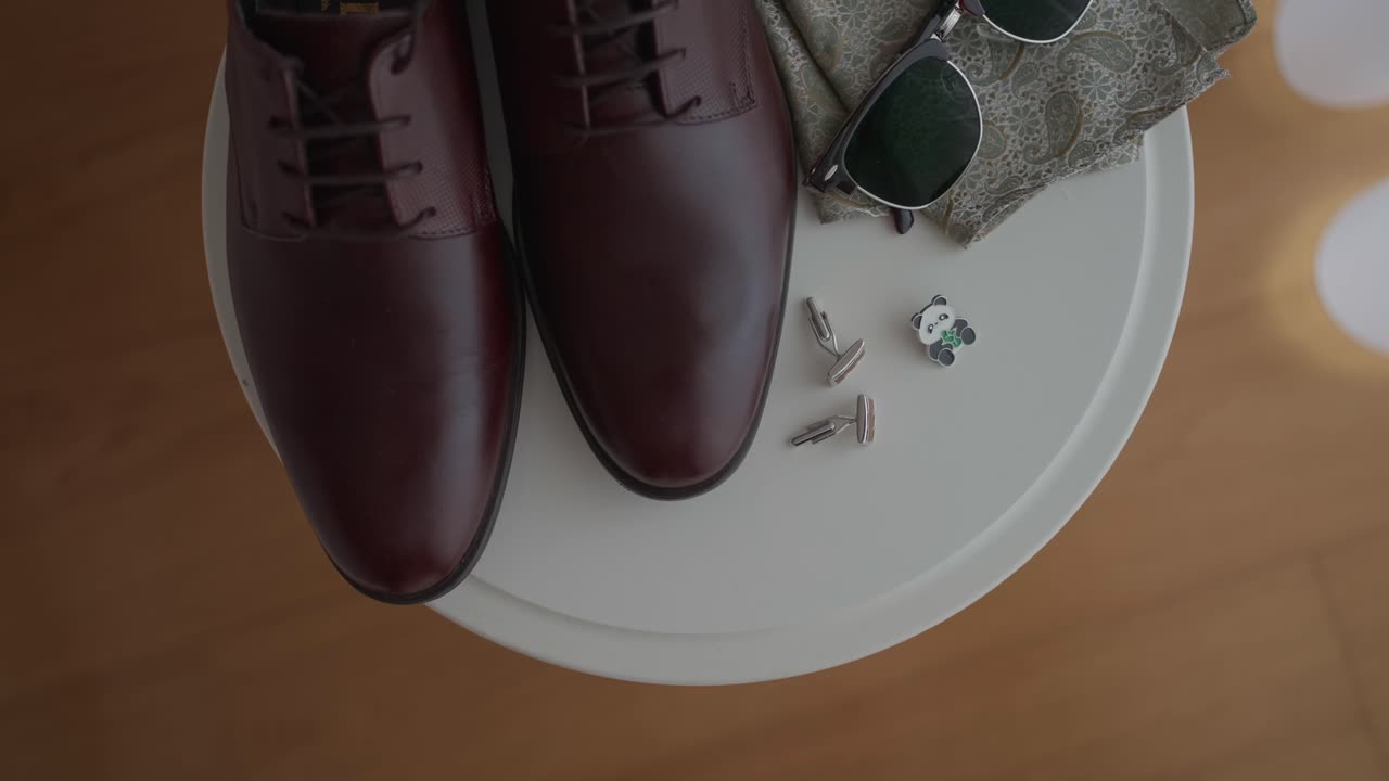 Top view of polished brown leather shoes, cufflinks, and sunglasses arranged neatly on a round table