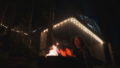 mujer de pie cerca de la hoguera calentando las manos. esposa envuelta en una cómoda manta se esfuerza por evitar el frío de la noche mientras está de pie en el patio de la casa de glamping