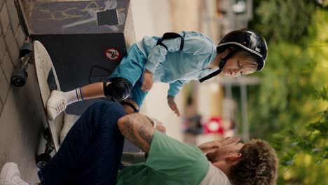 Vertical-video:-Little-blond-boy-in-blue-clothes-in-protective-clothing-for-riding-a-skateboard-in-a-black-helmet-high-fives-his-dad-with-curly-hair-in-a-Green-T-shirt-in-a-skate-park-in-summer