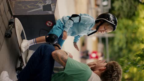video vertical: pequeño niño rubio con ropa azul en ropa de protección para montar una patineta en un casco negro high-fives a su padre con el cabello rizado en una camiseta verde en un parque de patinaje en verano