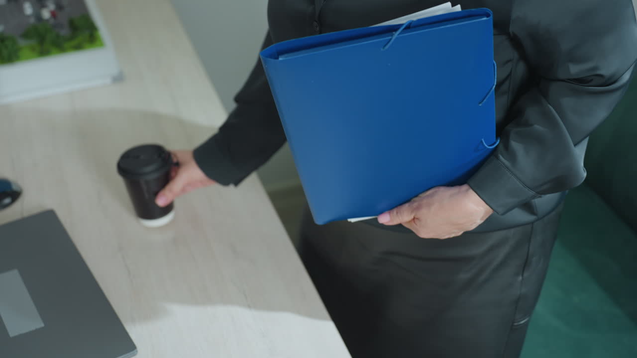Professional woman in black outfit places folder with documents and smartphone on desk beside coffee cup, then pulls laptop forward to begin remote office work in stylish modern workspace