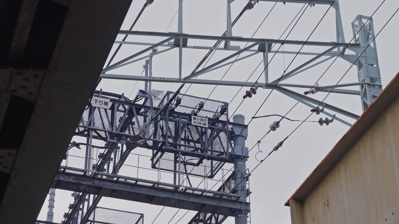 A static low-angle shot of intricate train power lines, insulators, and a steel gantry structure against an overcast sky at a Japanese station.
