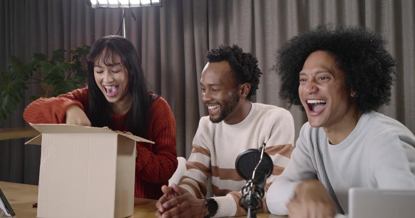 Diverse co-hosts unboxing cardboard box at studio table, inspecting black oval case for review